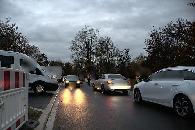 Einfahrt der Schule links, rechts die Grünanlage, aus beiden Richtungen fahren Autos dazwischen Lotsen, die Kinder über die Straße geleiten.