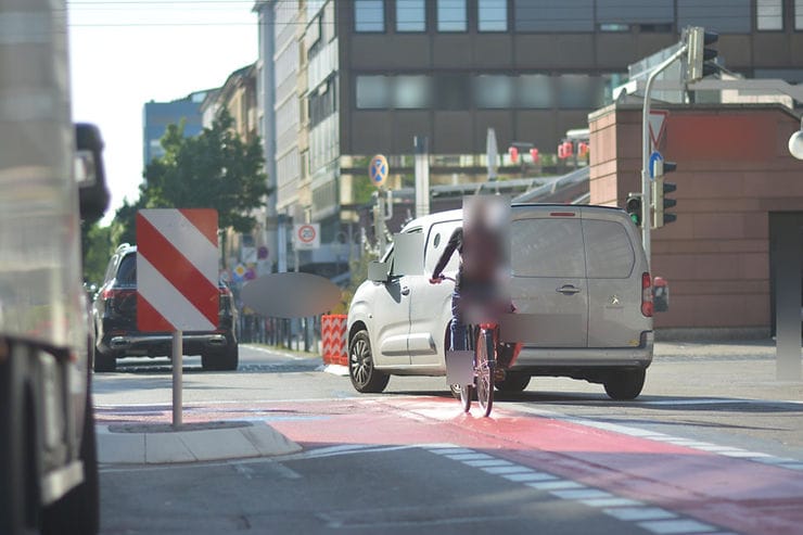 Absperrung recht, Absperrung links, Radweg aufgezeichnet dazwischen, Straße gut querbar, Autofahrer überholt Radfahrerin rechts und schneidet ihr den Weg ab