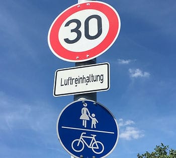 Tempo 30 Schild, darunter Schild Luftreinhaltung, darunter Schild geteilter Fußgänger- und Radweg, an einem Pfosten, vor blauem Himmel