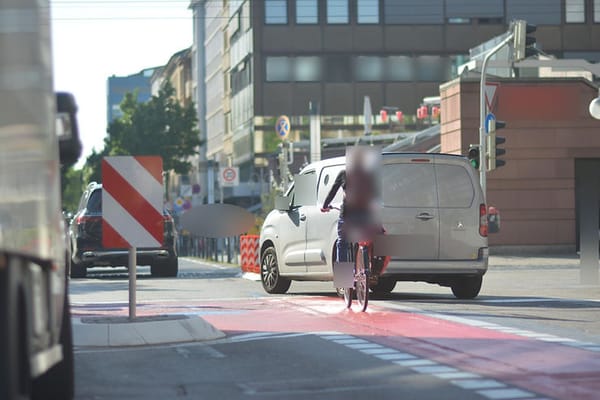 Absperrung recht, Absperrung links, Radweg aufgezeichnet dazwischen, Straße gut querbar, Autofahrer überholt Radfahrerin rechts und schneidet ihr den Weg ab